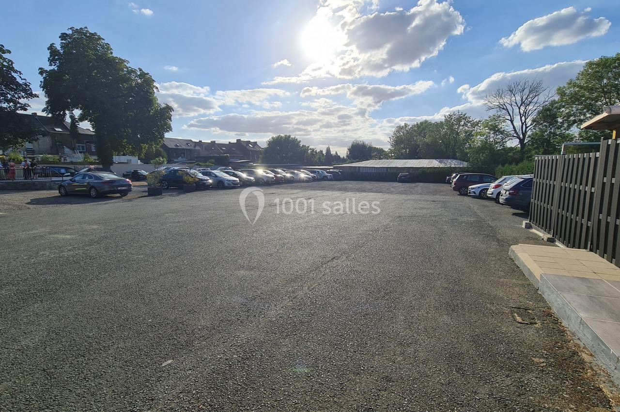 Parking extérieur en gravier avec plusieurs voitures stationnées, entouré d'arbres et de bâtiments sous un ciel ensoleillé.
