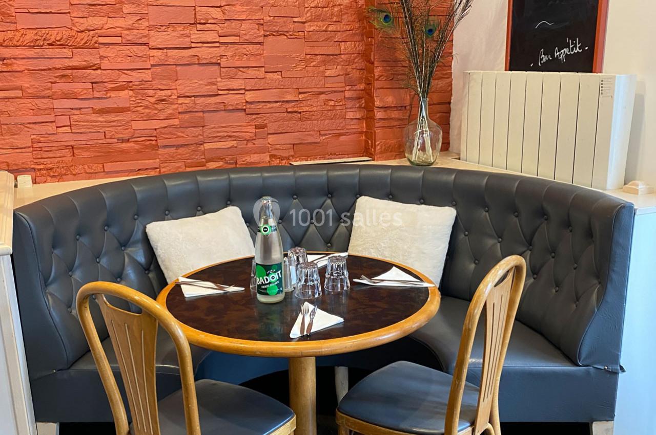 Banquette en cuir gris autour d'une table ronde en bois, avec chaises en bois, bouteille d'eau et couverts disposés.