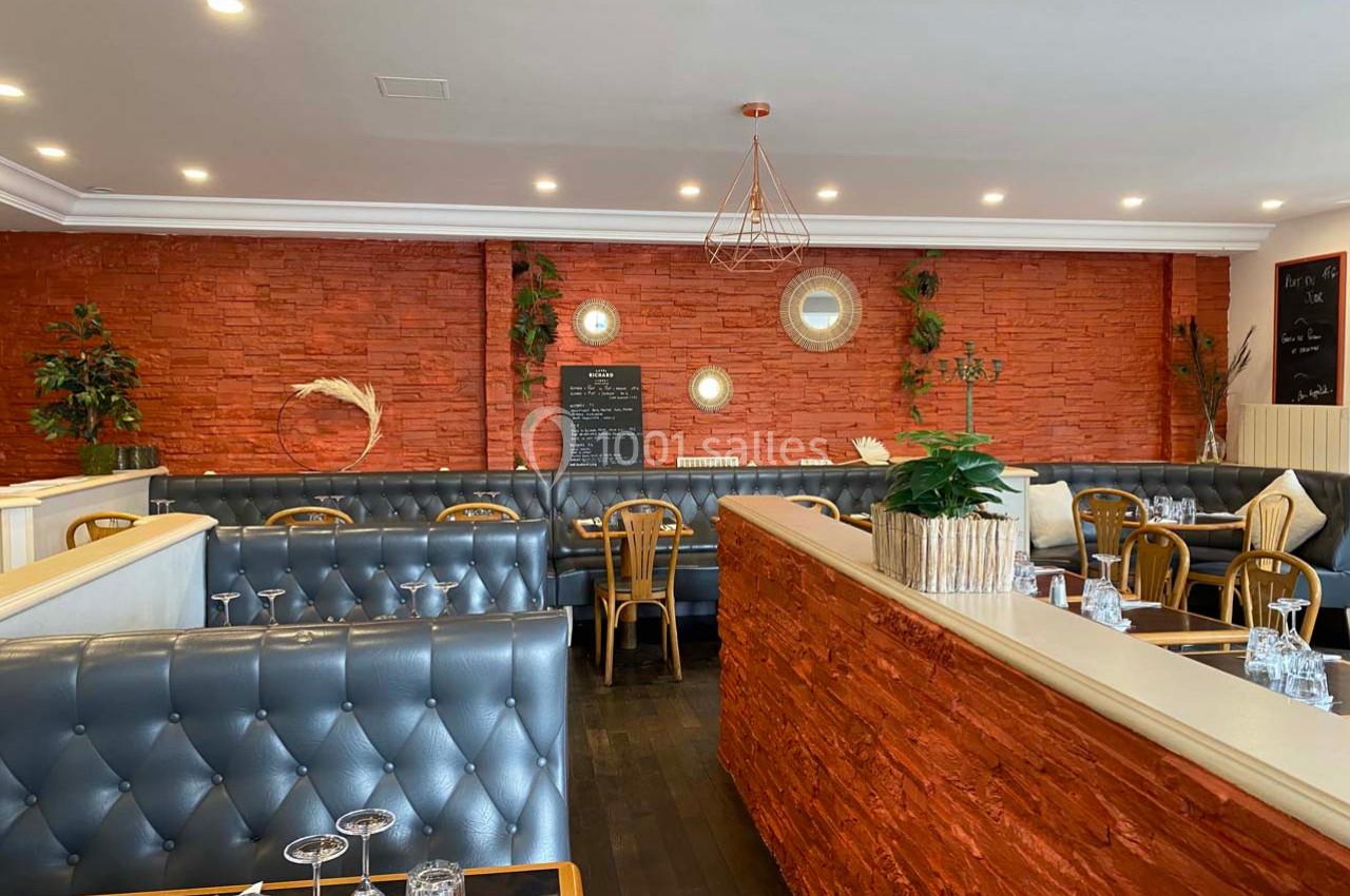 Salle de restaurant avec murs en briques rouges, banquettes en cuir gris, tables dressées et décoration végétale.