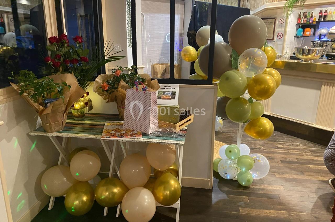 Décoration festive avec ballons dorés et verts, bouquets de fleurs et cadeaux sur une table en bois dans un espace intérieur.