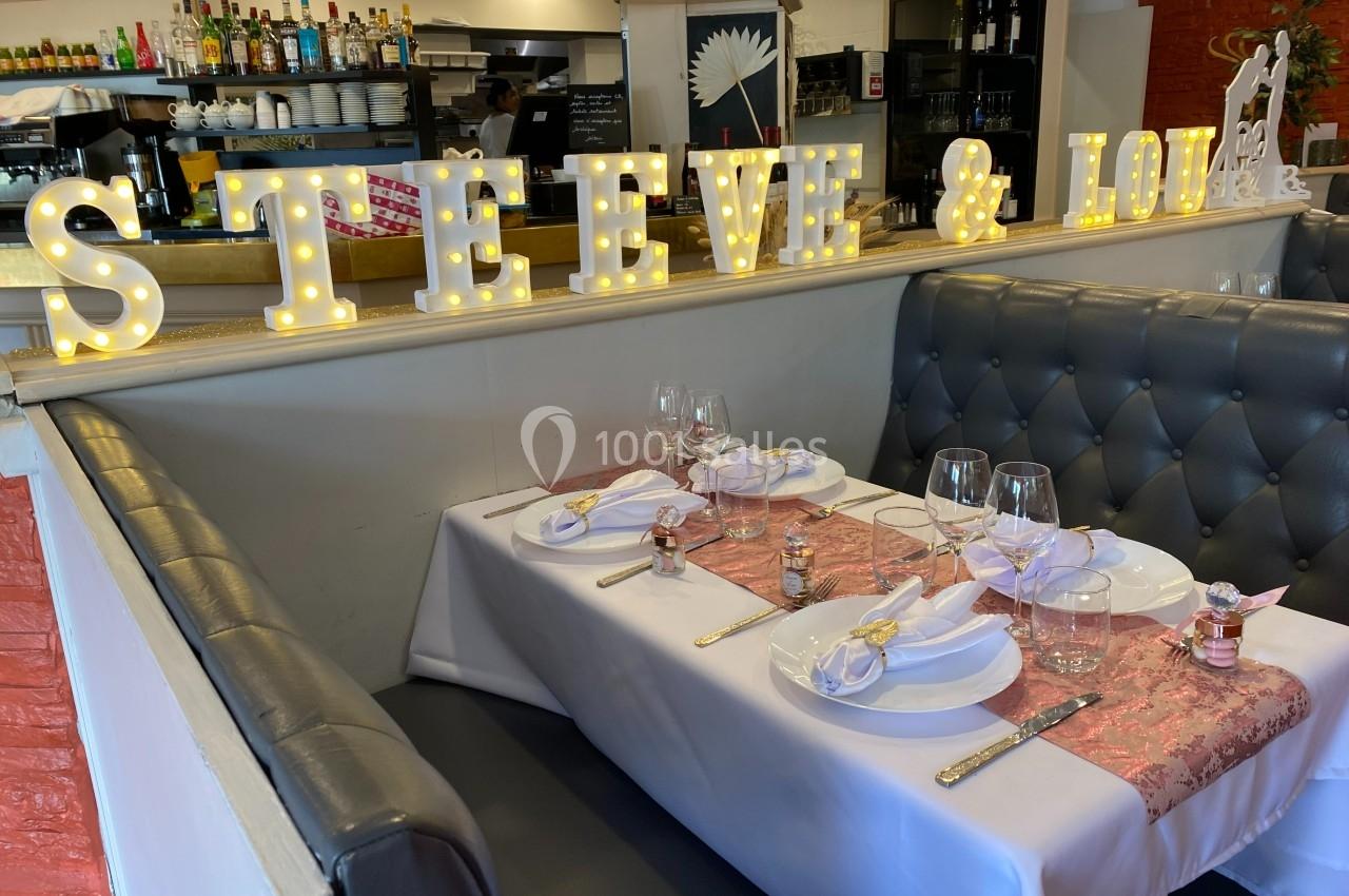 Table de restaurant décorée pour un événement, avec nappes blanches, vaisselle élégante et lettres lumineuses en arrière…