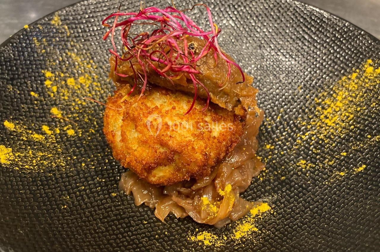 Croquette dorée servie sur un lit d'oignons caramélisés, garnie de pousses rouges et d'épices jaunes.