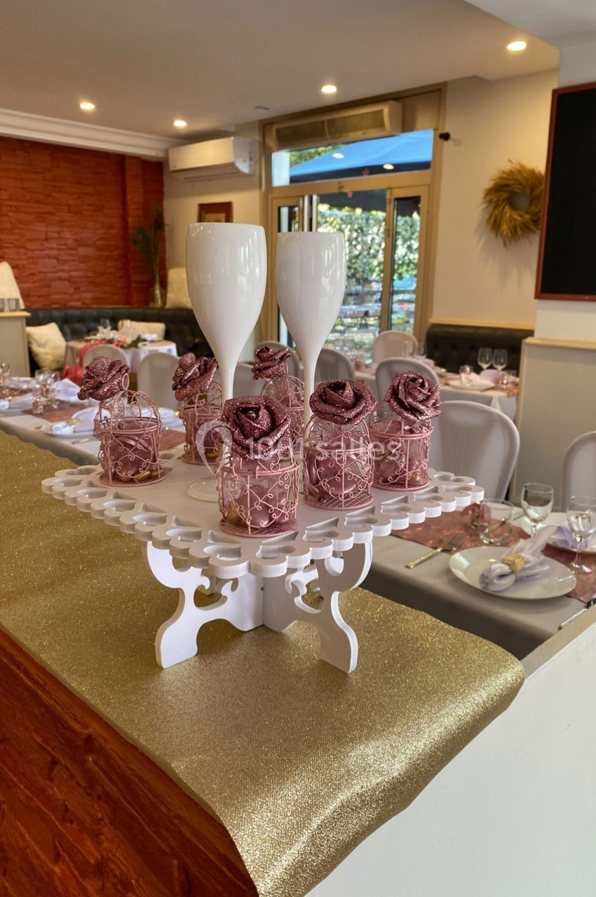 Présentoir décoratif avec roses artificielles roses et flûtes blanches, dans une salle de réception élégamment dressée.