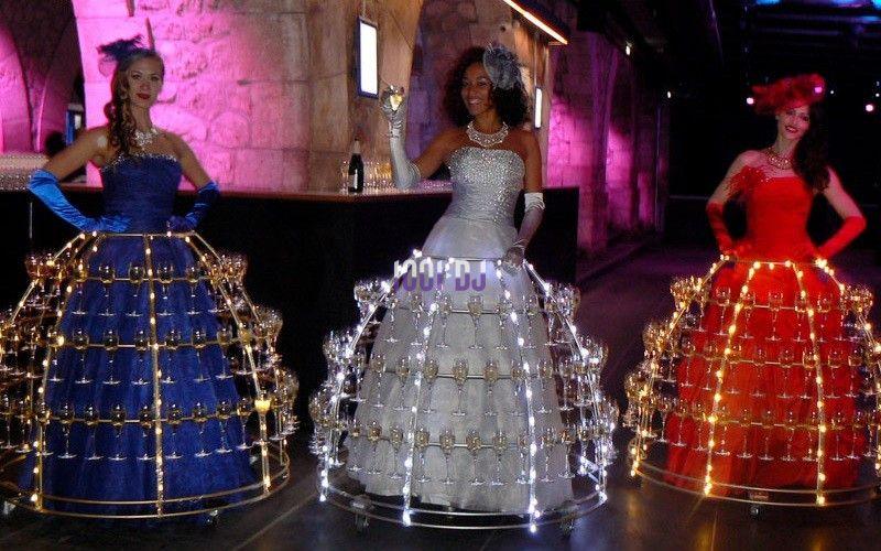 Trois femmes en robes lumineuses portant des structures métalliques garnies de verres à champagne dans un décor festif.