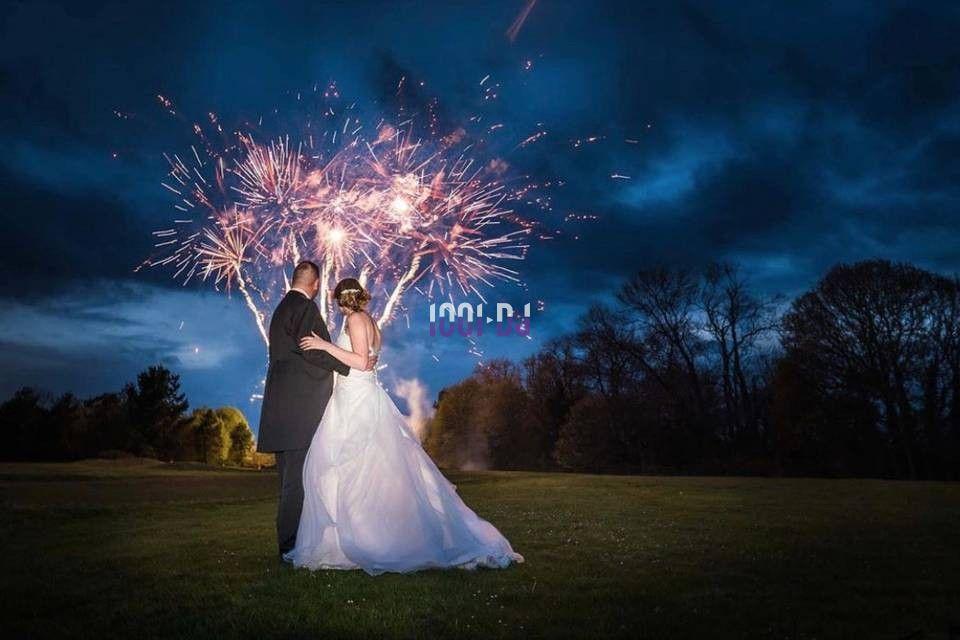 Un couple en tenue de mariage regarde un feu d'artifice illuminant un ciel nocturne nuageux.