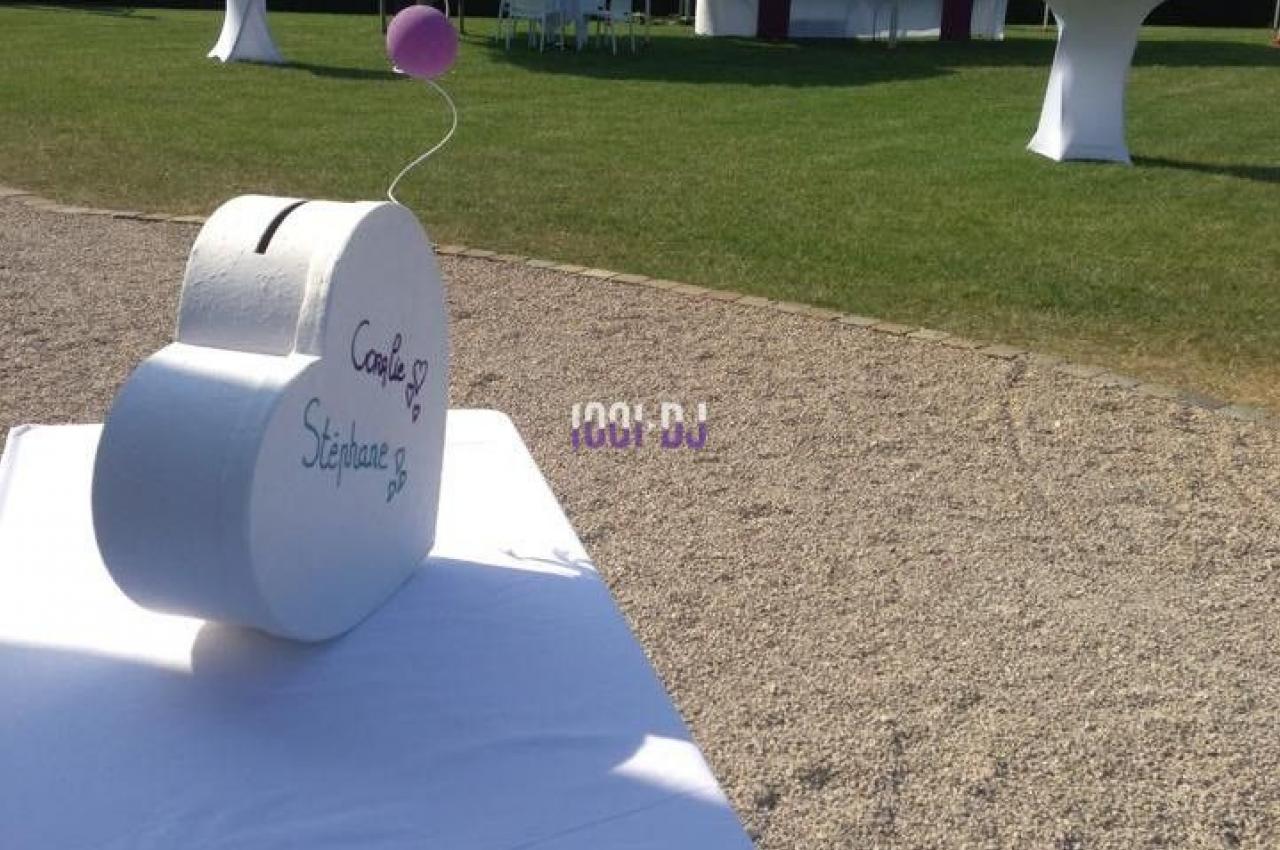 Urne de mariage en forme de cœur sur une table blanche, avec des ballons et un espace de réception en arrière-plan.