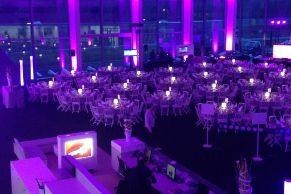 Salle de réception éclairée en violet, avec des tables rondes dressées et des chaises blanches, vue depuis une mezzanine.