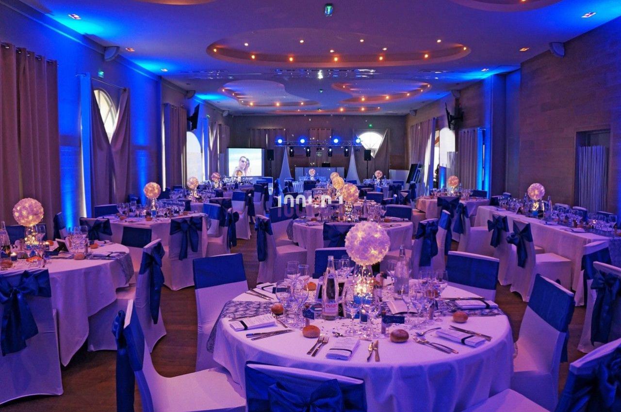 Salle de réception décorée avec des tables rondes, nappes blanches, chaises ornées de nœuds bleus et éclairage tamisé.