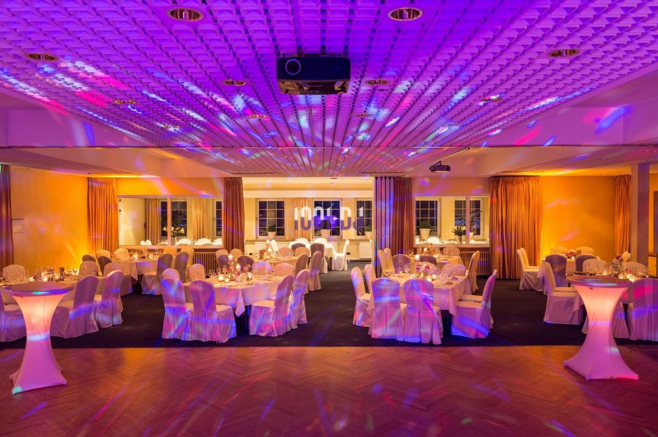 Salle de réception éclairée par des lumières colorées, avec tables dressées et chaises recouvertes de housses blanches.