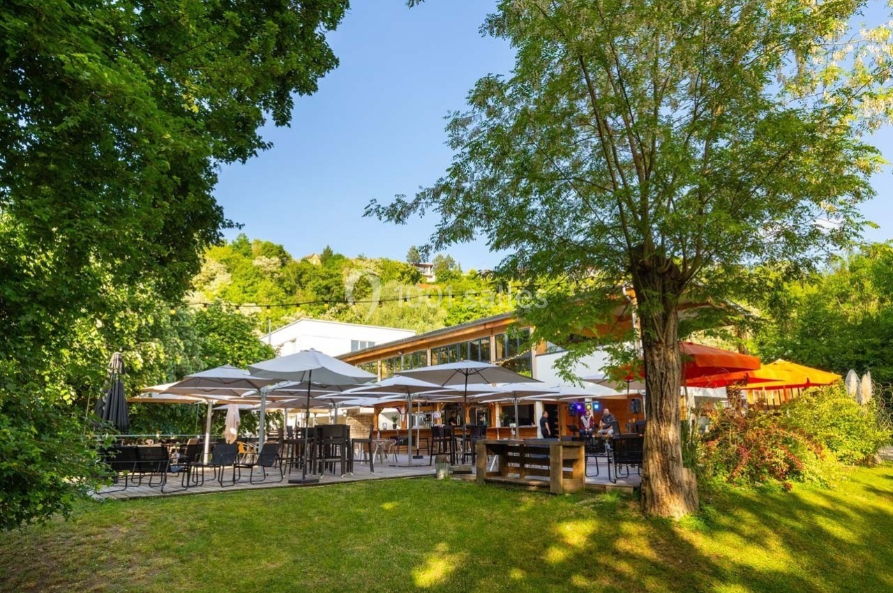 Terrasse d'un restaurant entourée de verdure, avec des tables, des parasols et un bâtiment moderne en arrière-plan.