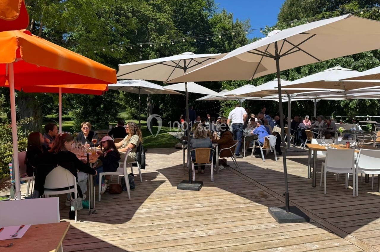 Terrasse en bois avec des tables et chaises occupées par des personnes, sous des parasols, entourée de verdure.