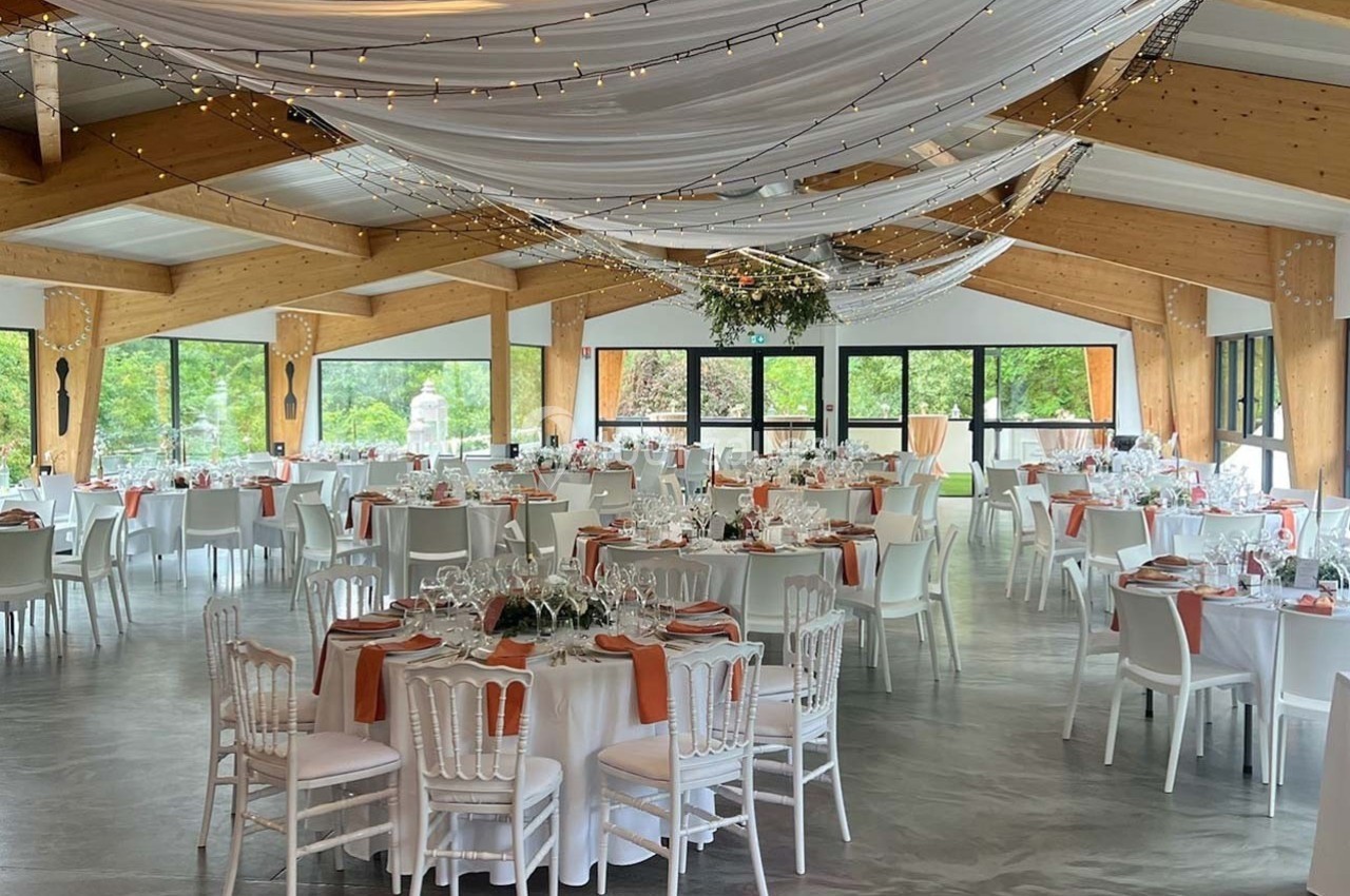 Salle de réception décorée avec des tables rondes dressées, nappes blanches, chaises blanches et guirlandes lumineuses…