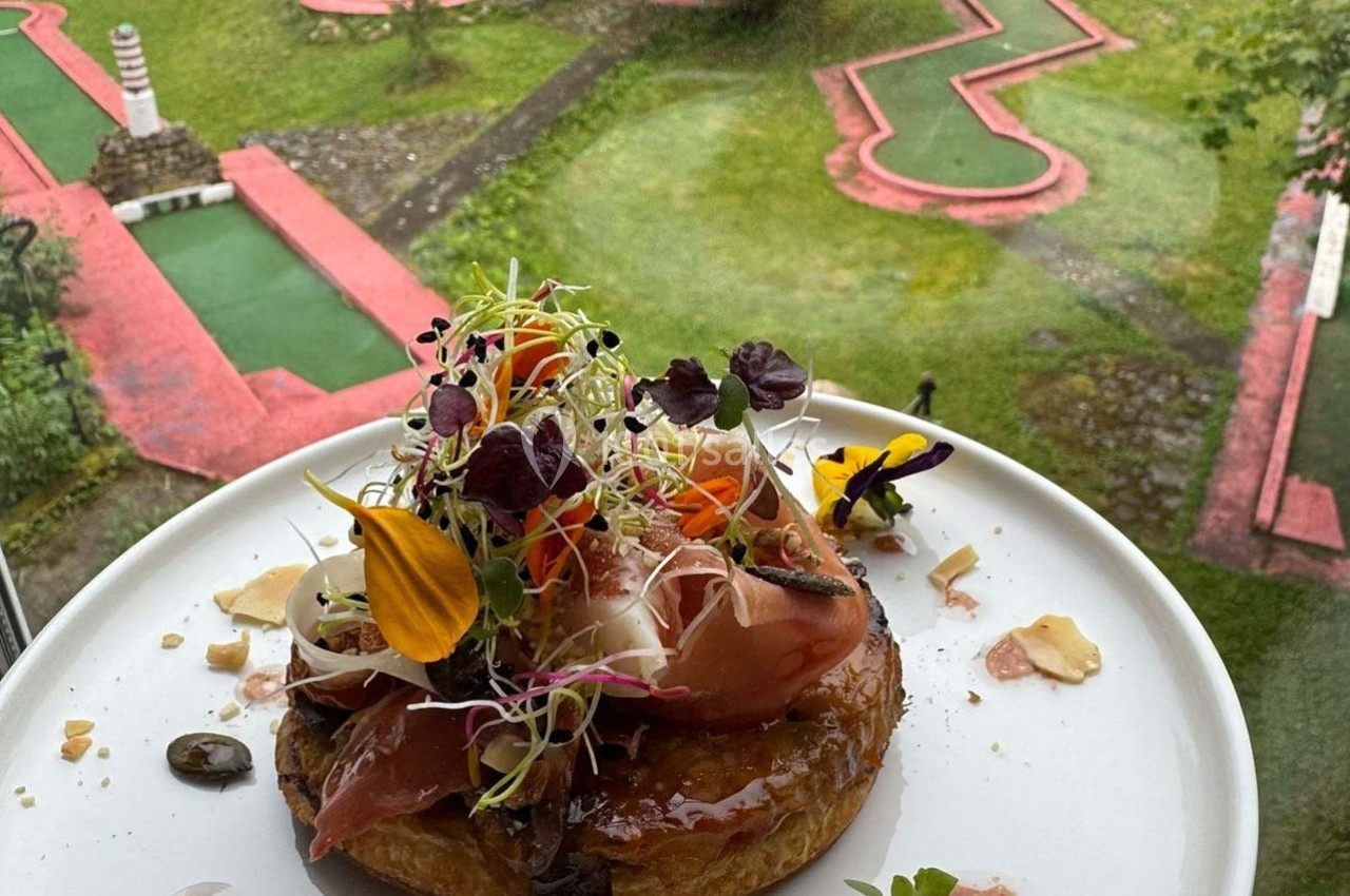 Tarte garnie de jambon, légumes, pousses et fleurs comestibles, avec un mini-golf en arrière-plan.
