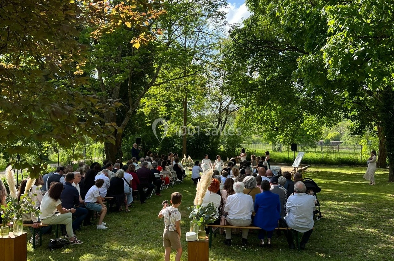 Cérémonie en plein air avec des invités assis sur des bancs dans un jardin verdoyant sous un ciel ensoleillé.