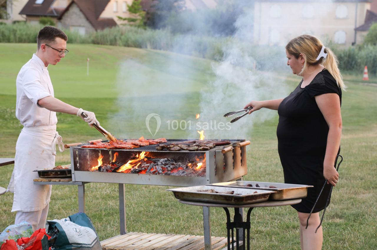 Un homme et une femme cuisinent des viandes et légumes sur un barbecue en plein air, dans un espace verdoyant.