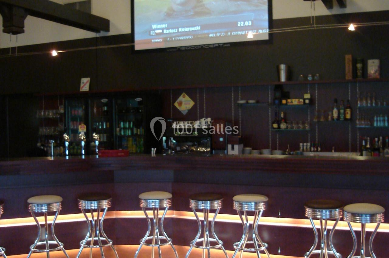 Un bar avec des tabourets alignés, un comptoir éclairé et un écran diffusant une émission au mur.