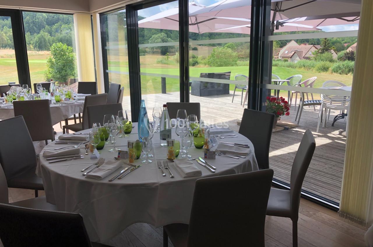 Salle de restaurant lumineuse avec tables dressées, vue sur une terrasse et un paysage verdoyant à travers de grandes baies…