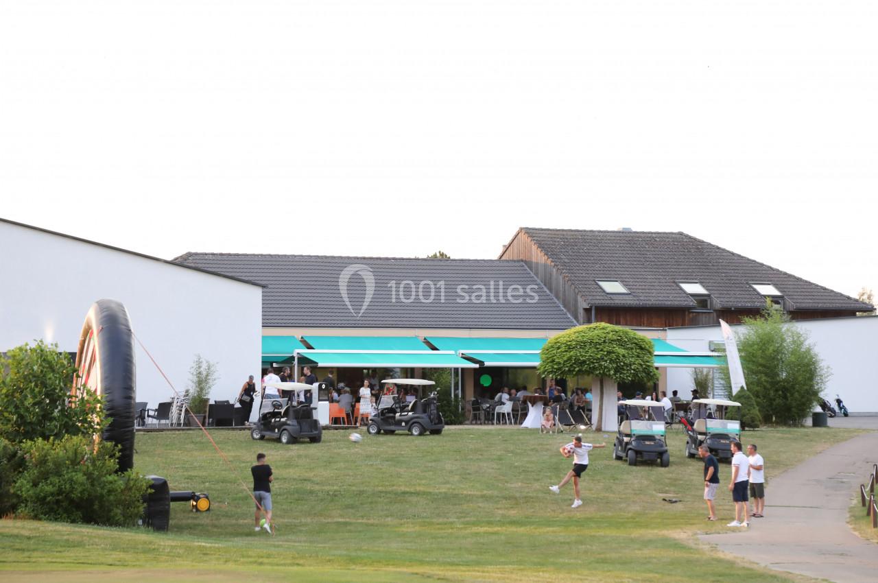 Des personnes rassemblées devant un bâtiment moderne avec des terrasses, des parasols et des voiturettes de golf.