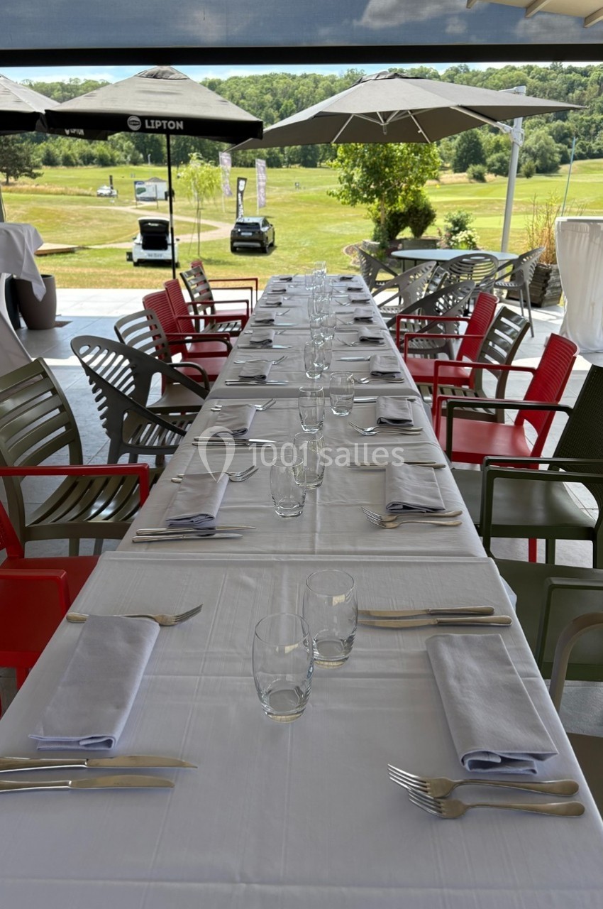 Table dressée avec nappes blanches, couverts et verres, sur une terrasse avec vue sur un paysage verdoyant.
