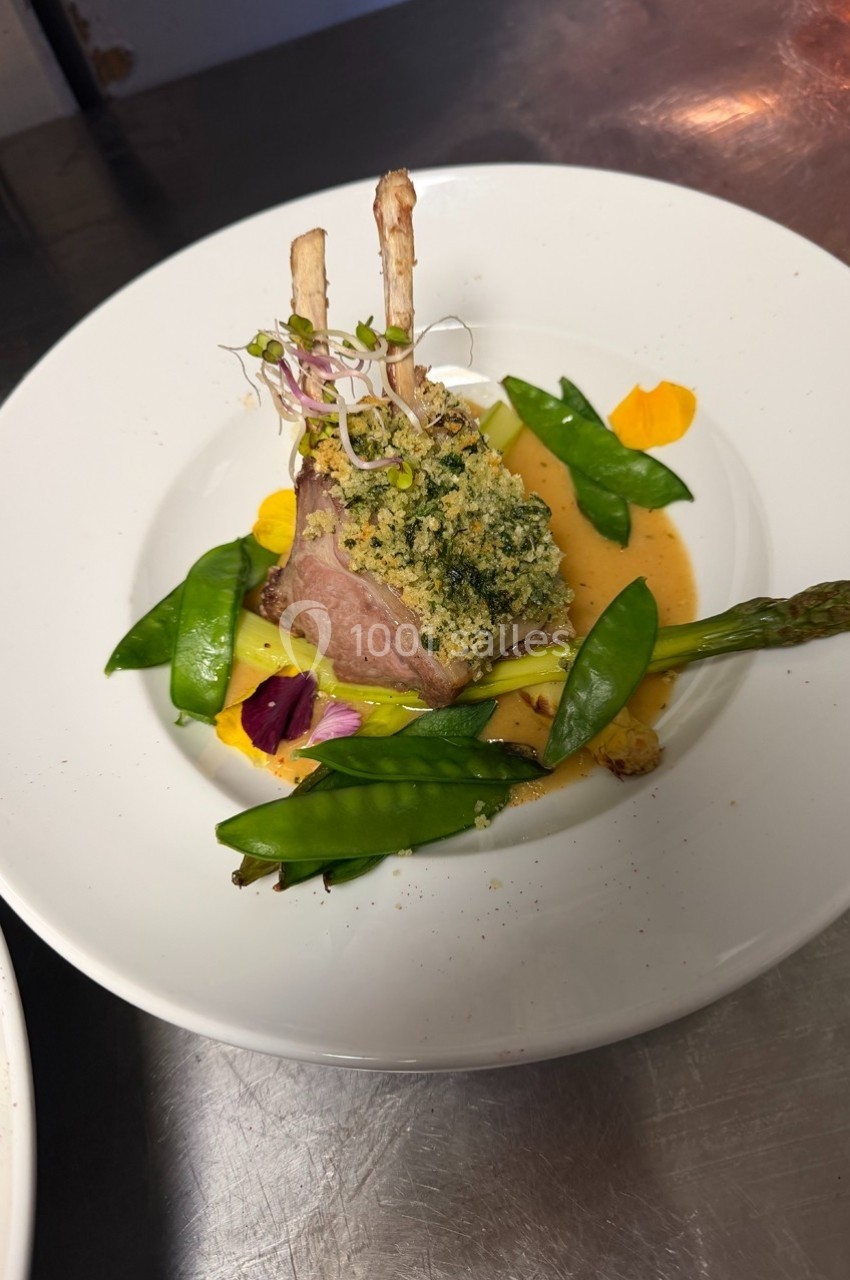 Carré d'agneau en croûte d'herbes, accompagné de légumes verts et d'une sauce dressée sur une assiette blanche.