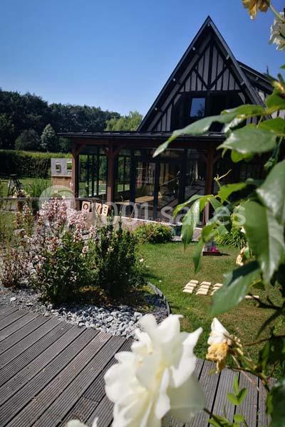 Jardin fleuri avec terrasse en bois, devant une maison à colombages et une véranda vitrée sous un ciel bleu.