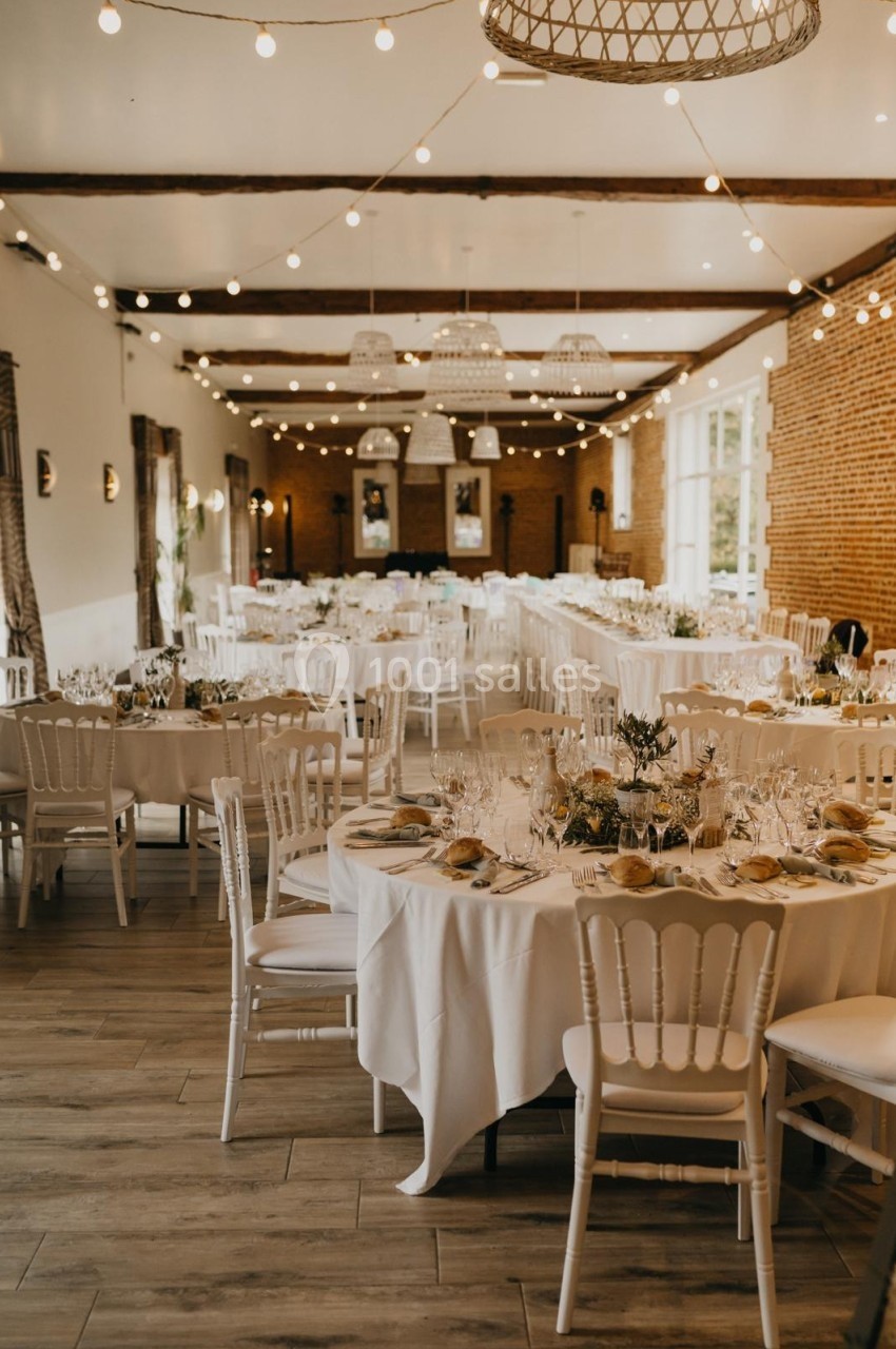 Salle de réception décorée avec des tables rondes dressées, chaises blanches et guirlandes lumineuses suspendues.