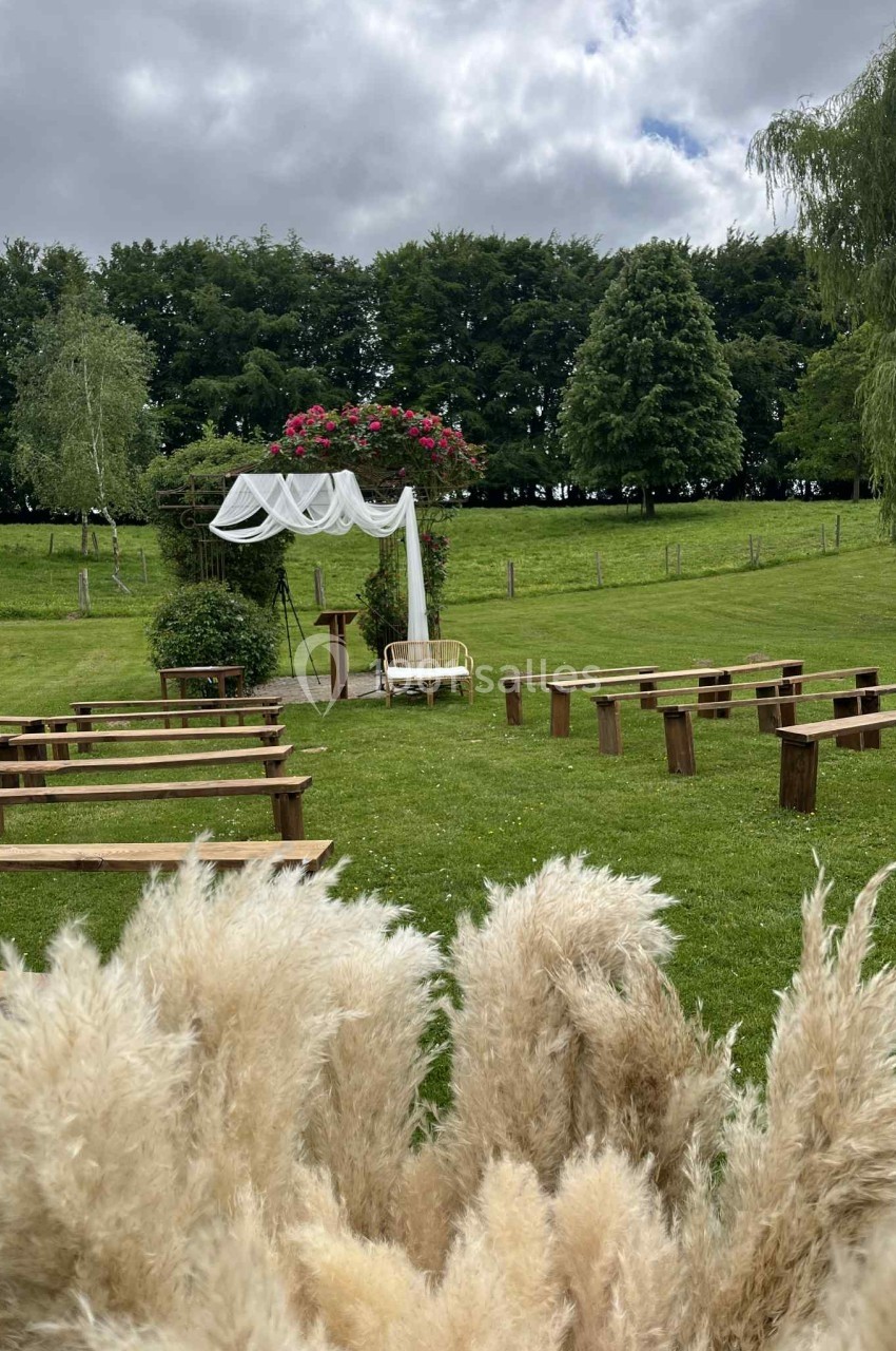 Vue d'une cérémonie en plein air avec arche décorée, bancs en bois et herbes de la pampa au premier plan.