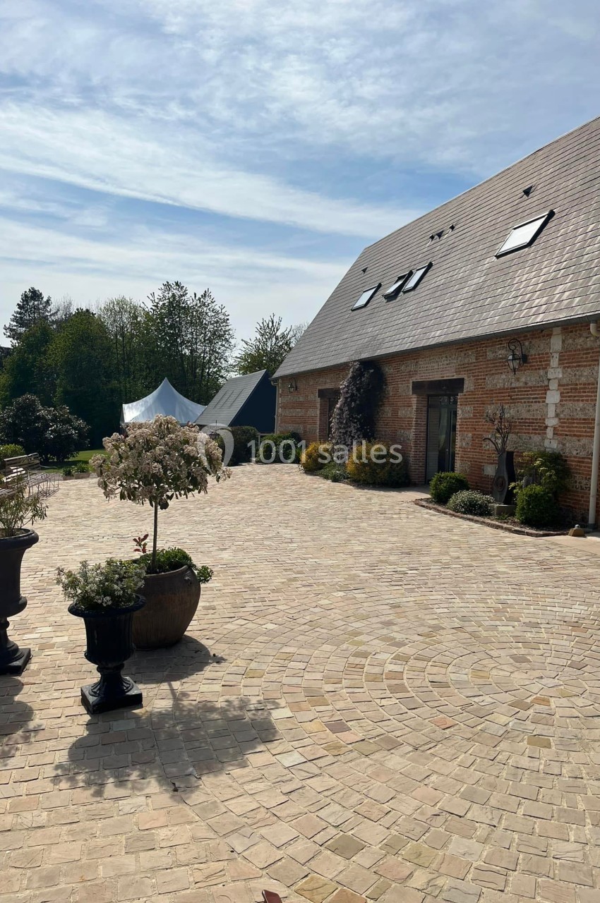 Cour pavée devant une maison en briques avec toit en ardoise, entourée de plantes et d'arbres sous un ciel dégagé.