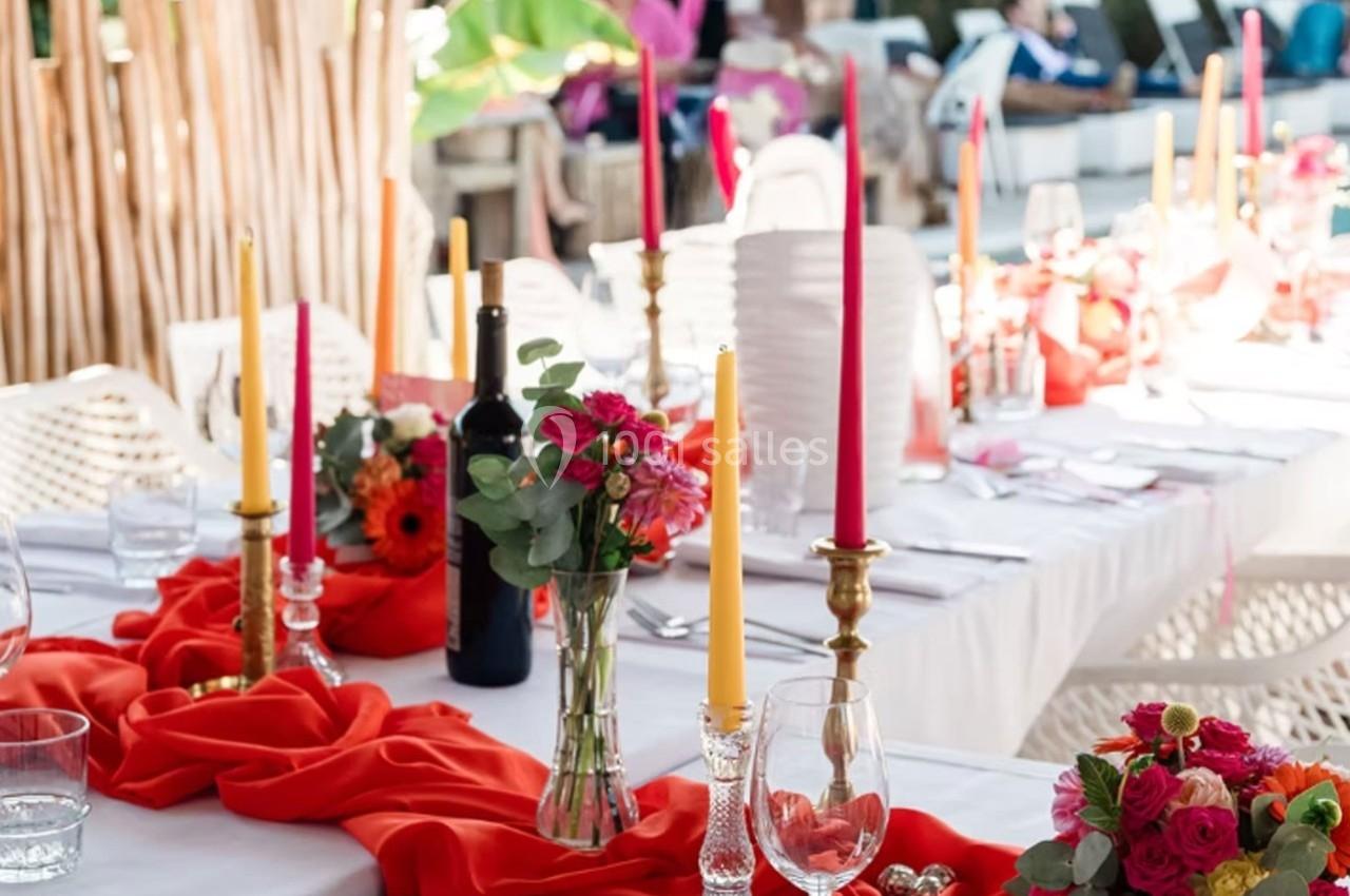 Table décorée avec des nappes rouges, chandeliers, bougies colorées, fleurs et une bouteille de vin dans un cadre extérieur.