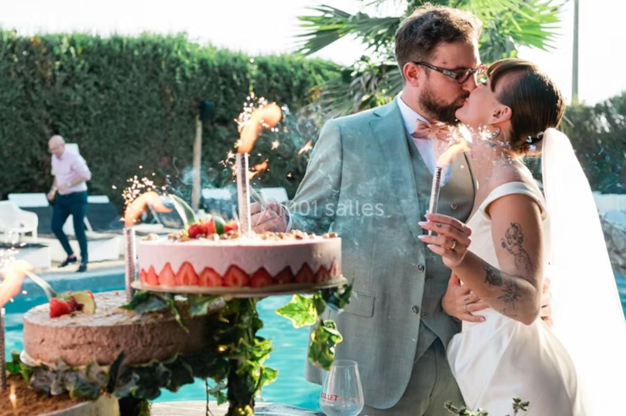 Un couple de mariés s'embrasse près d'une piscine, entouré de gâteaux décorés et de cierges magiques allumés.