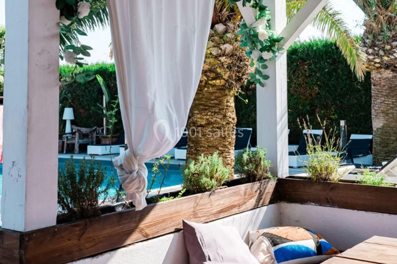 Terrasse ombragée avec rideaux blancs, coussins colorés, plantes en jardinières et vue sur une piscine entourée de palmiers.
