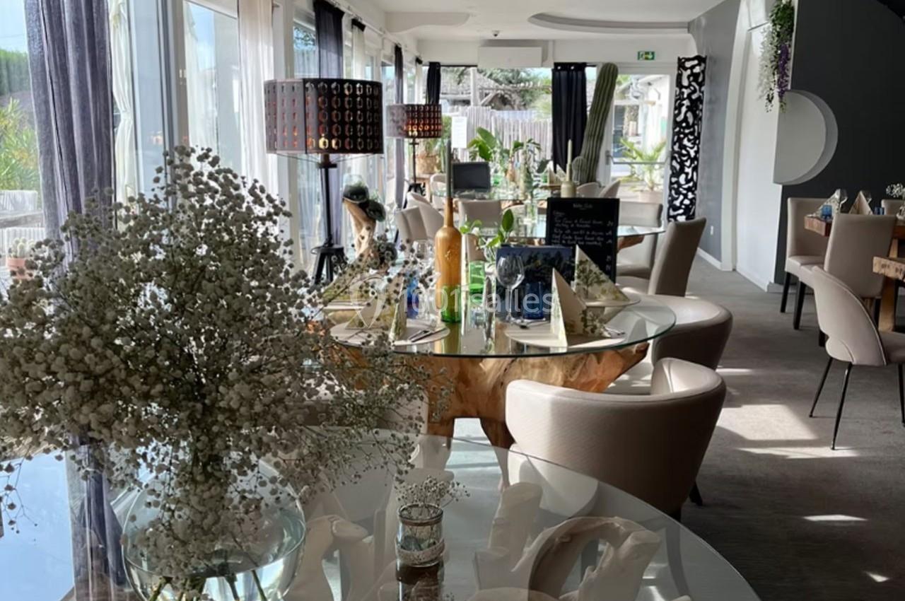 Salle lumineuse avec tables en verre, chaises beige, décorations florales et grandes baies vitrées donnant sur l'extérieur.