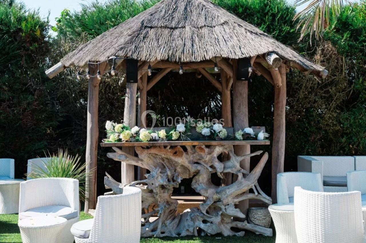 Kiosque en bois avec toit en chaume, décoré de fleurs blanches, entouré de fauteuils blancs sur une pelouse.