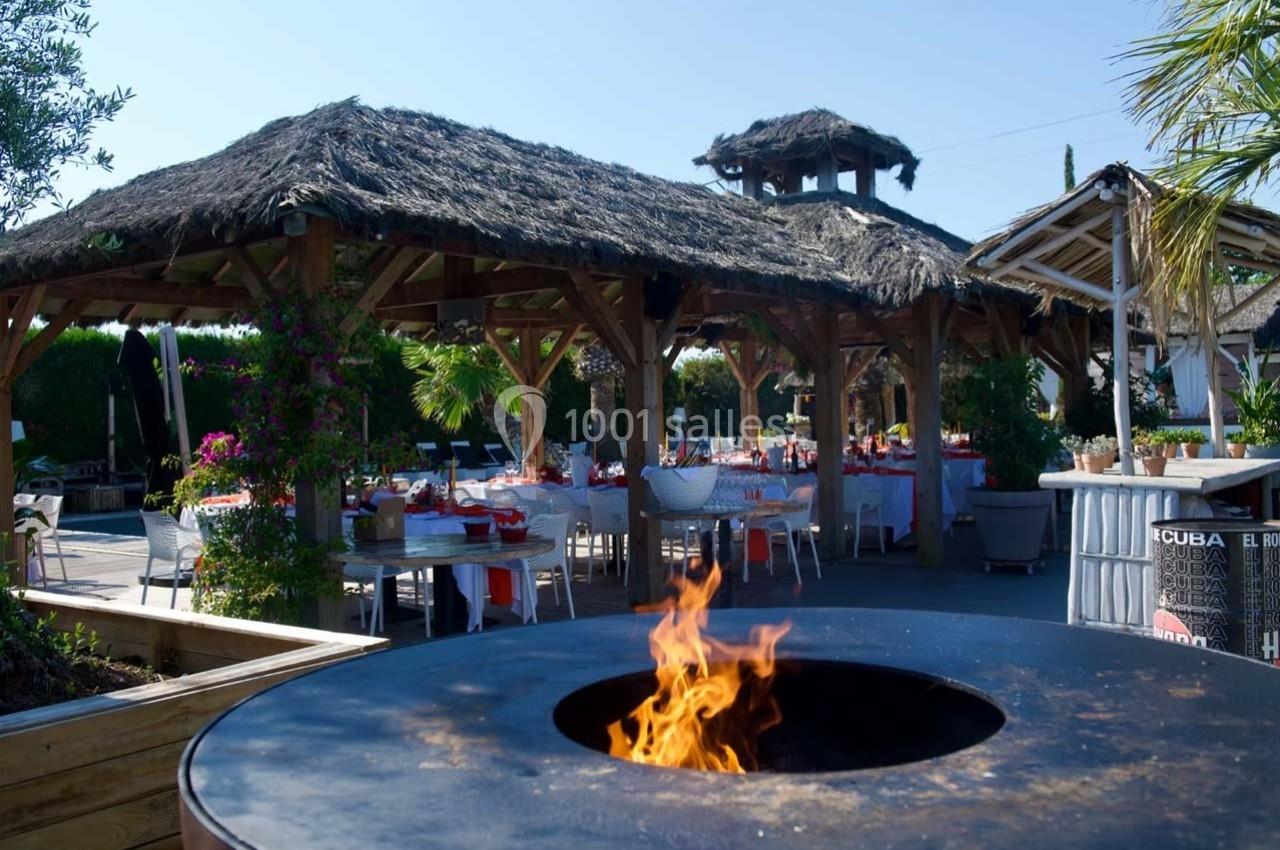 Terrasse extérieure avec tables dressées sous des toits en chaume, et un brasero allumé au premier plan.