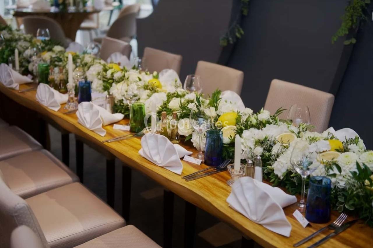 Table en bois décorée de fleurs blanches et jaunes, serviettes pliées, verres et couverts pour un repas formel.