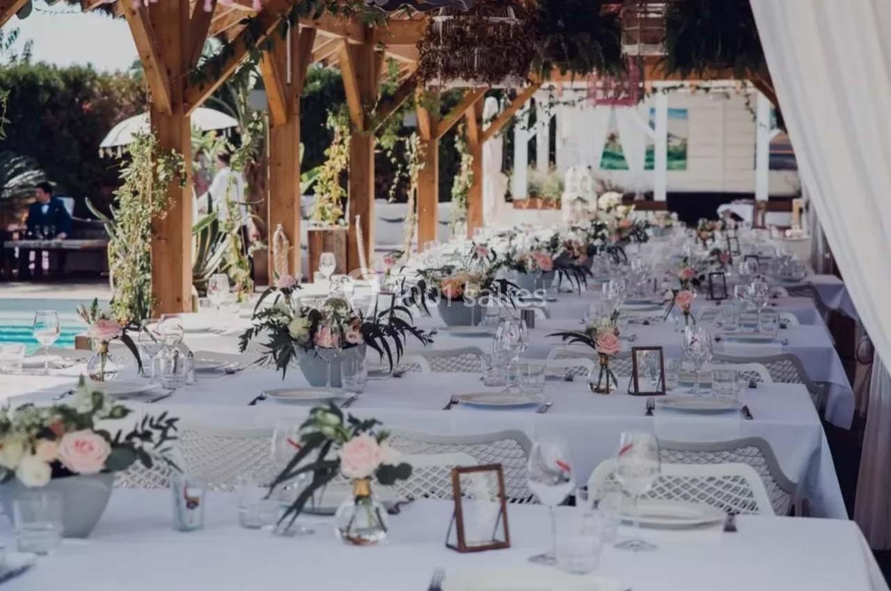 Tables décorées avec des fleurs et nappes blanches sous une pergola en bois, prêtes pour un événement en extérieur.
