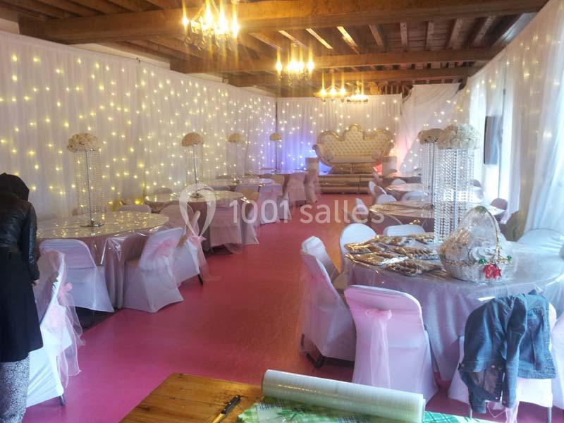 Salle décorée pour un événement, avec tables rondes, nappes blanches, guirlandes lumineuses et fauteuil en arrière-plan.