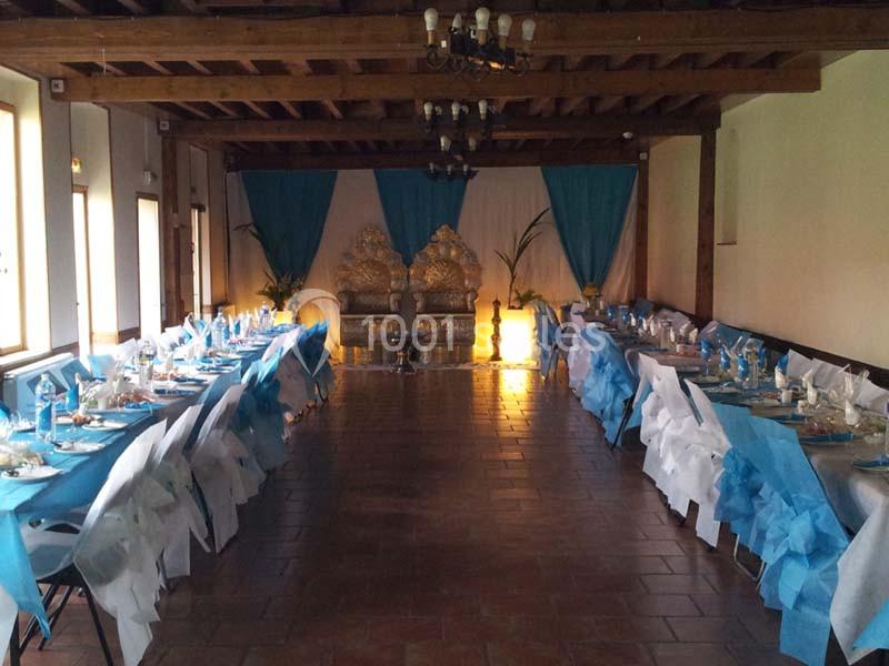 Salle de réception décorée avec des tables alignées, nappes blanches et accents bleus, et un trône doré en fond.