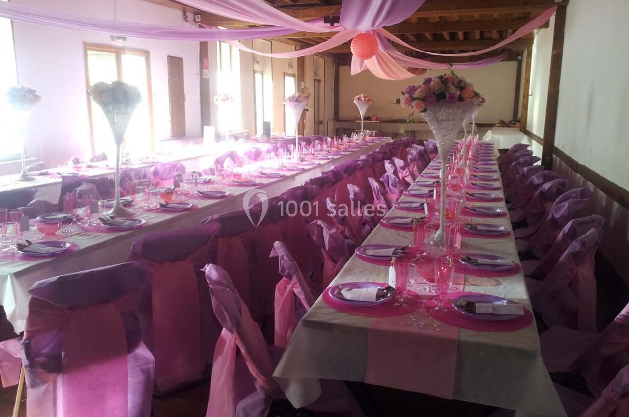 Salle décorée pour un événement avec des tables alignées, nappes roses, chaises assorties et centres de table fleuris.