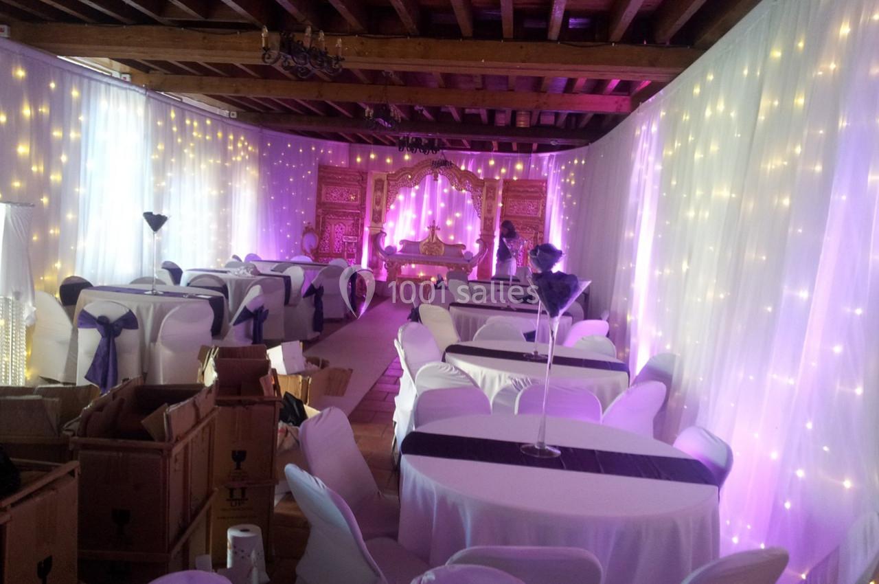 Salle décorée pour un événement avec des tables drapées de blanc, des lumières féeriques et un espace central aménagé.