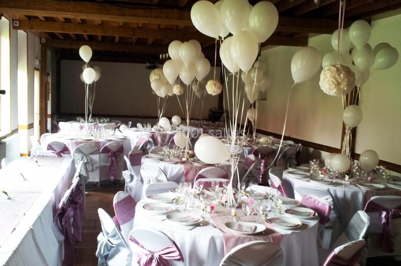 Salle décorée pour une réception avec des tables dressées, nappes blanches, rubans roses et ballons blancs.