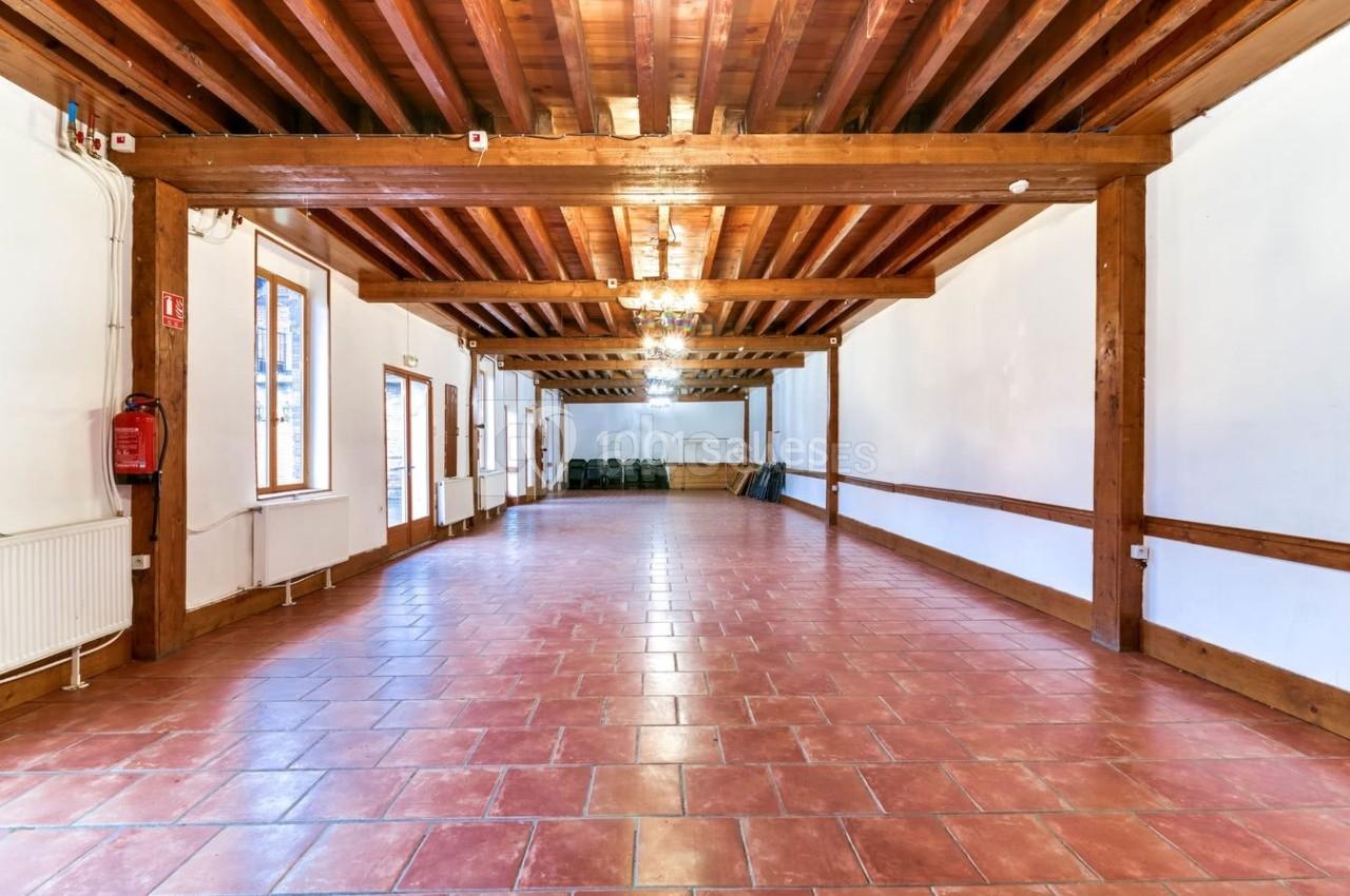 Salle vide avec sol en carrelage rouge, murs blancs, poutres en bois au plafond et grandes fenêtres laissant entrer la…