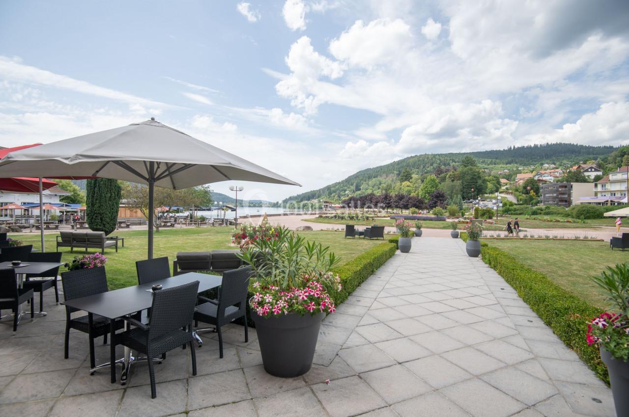 Terrasse avec tables et chaises noires, bordée de plantes fleuries, donnant sur un parc et un paysage vallonné.