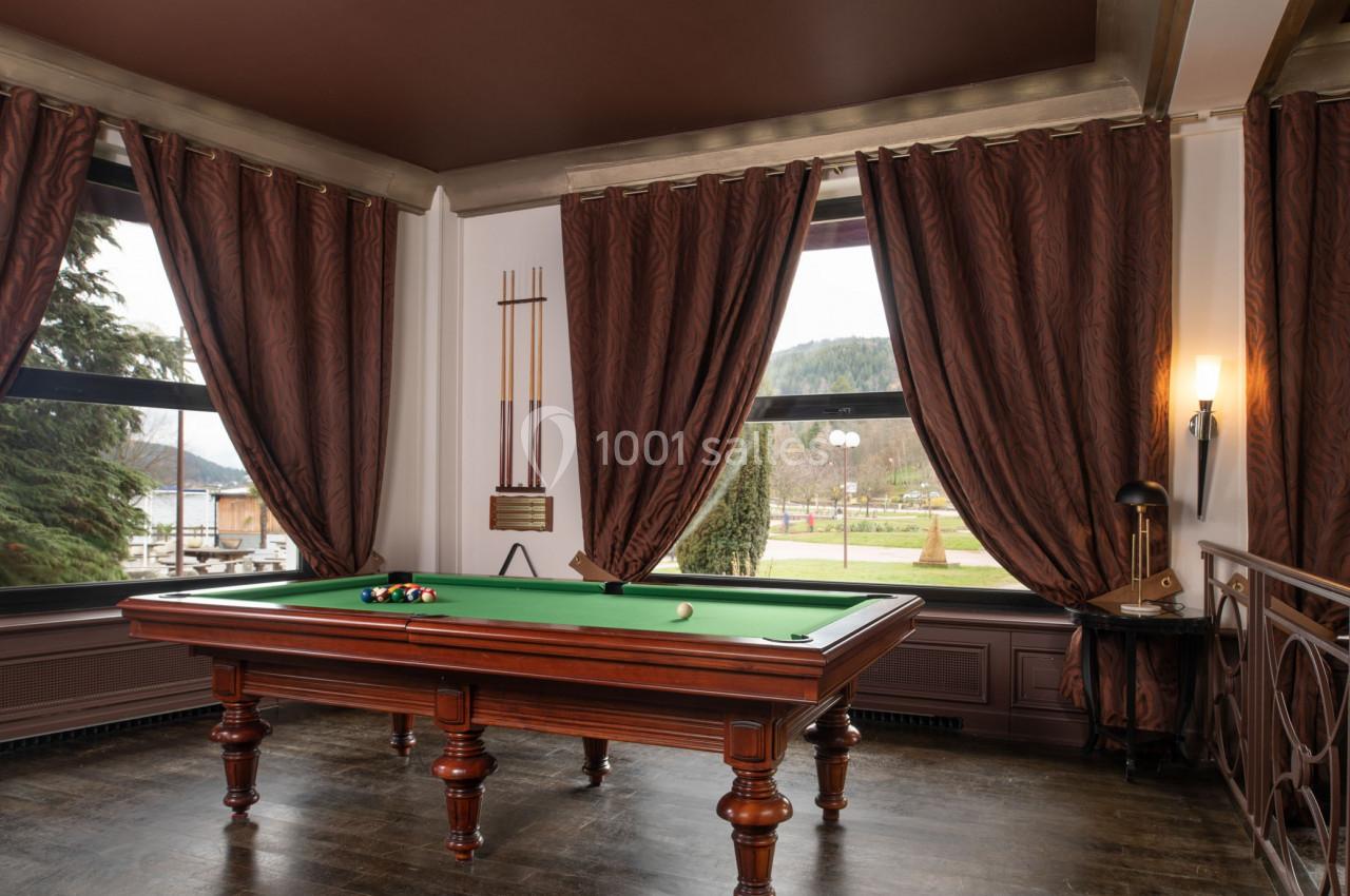 Table de billard en bois avec tapis vert dans une pièce élégante aux rideaux marron, vue sur un parc extérieur.