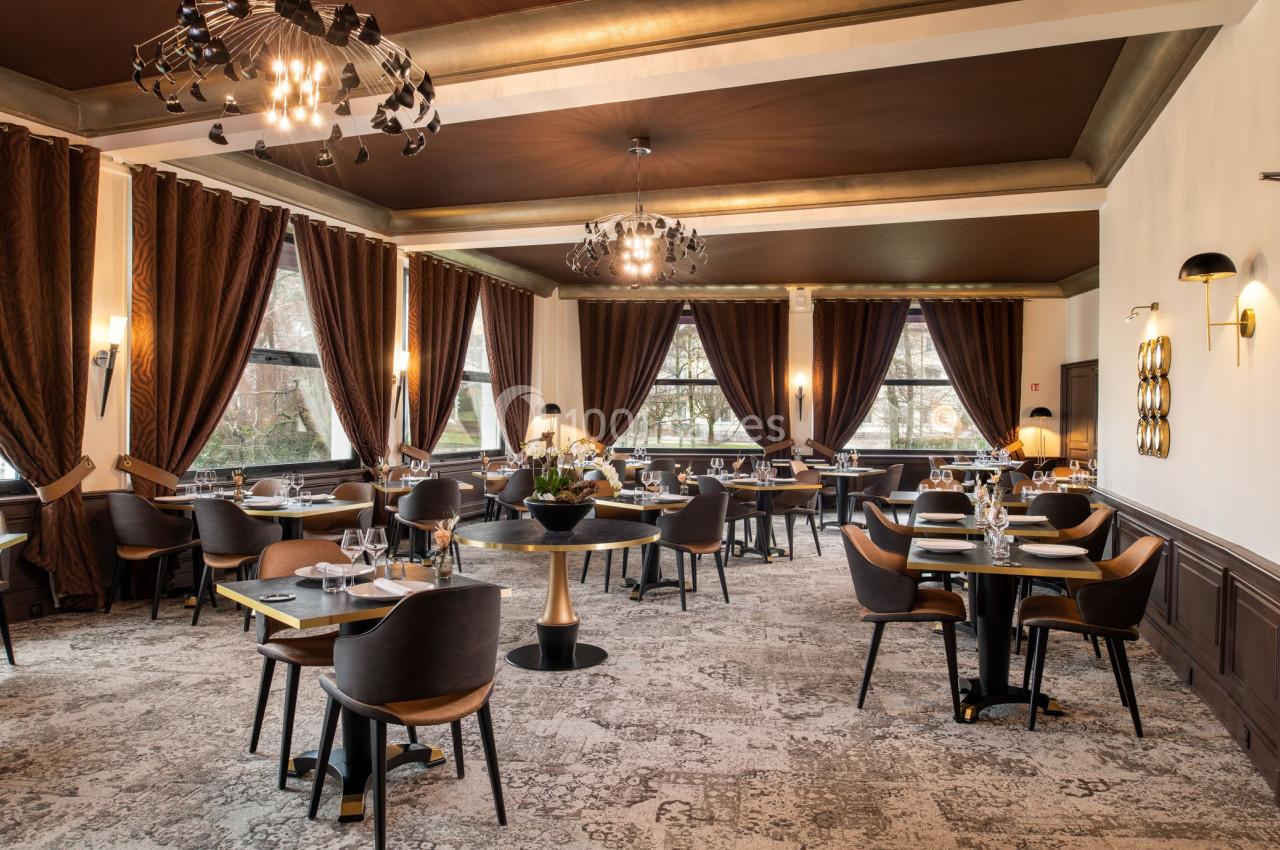 Salle de restaurant élégante avec tables dressées, chaises modernes, grandes fenêtres et rideaux sombres.