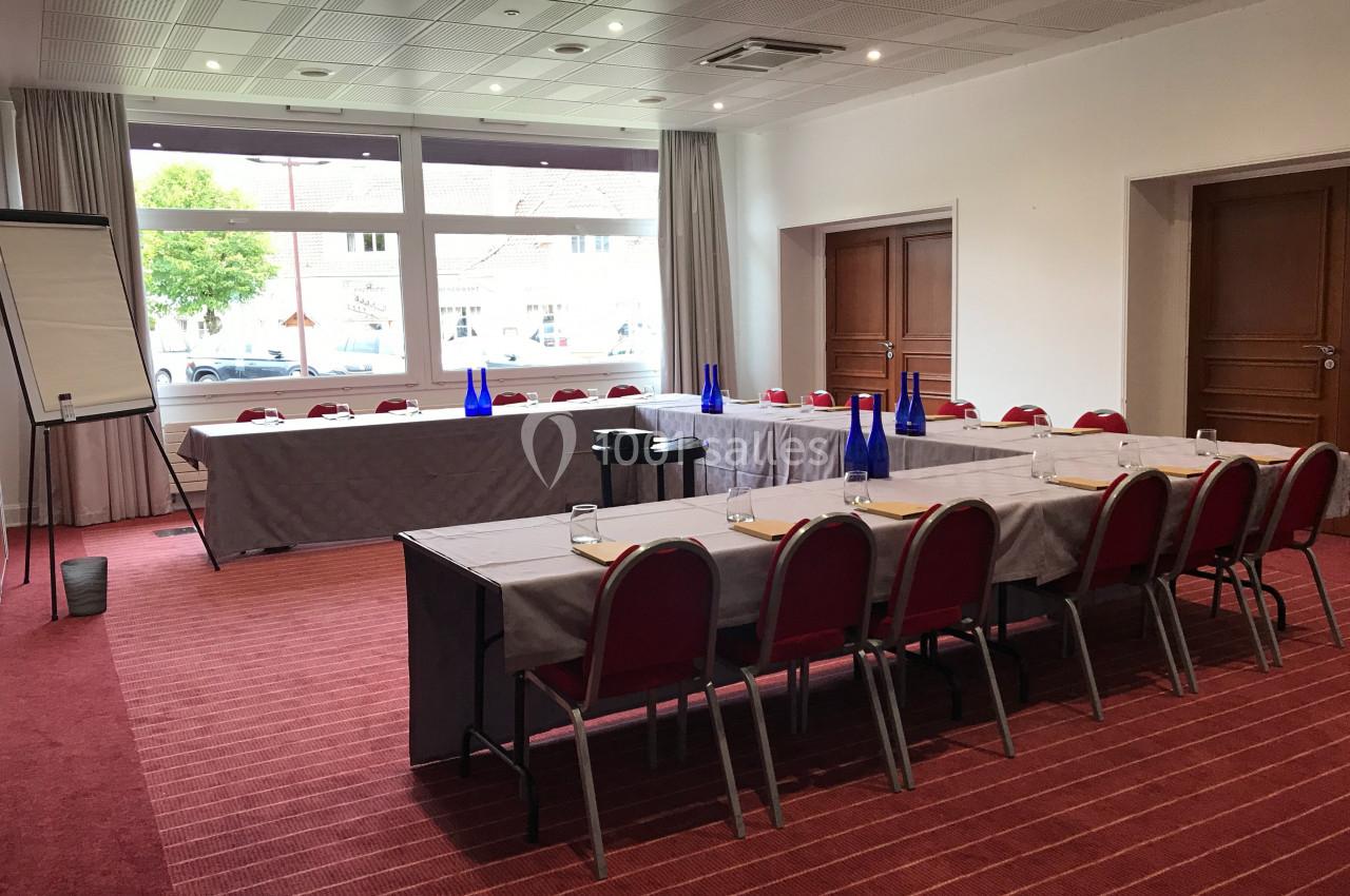 Salle de réunion lumineuse avec tables disposées en U, chaises rouges, tableau de conférence et bouteilles d'eau bleues.