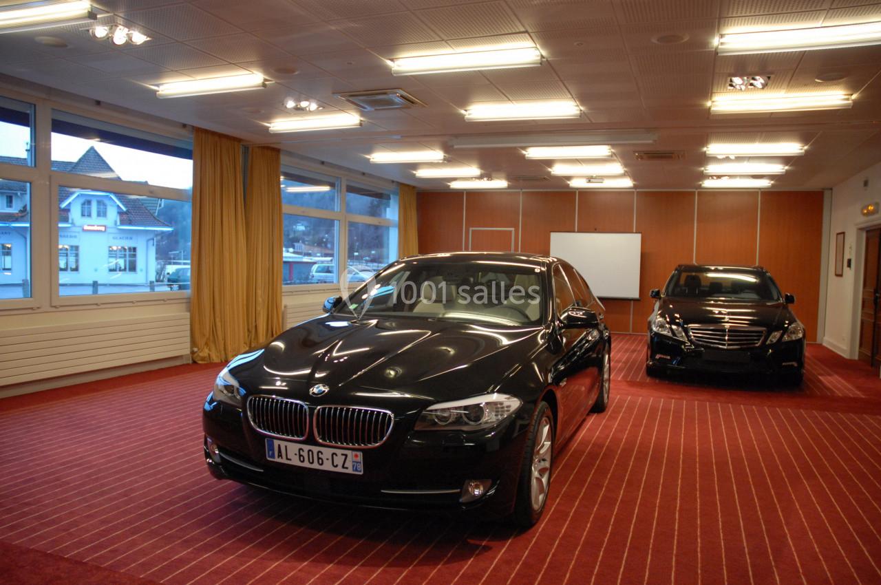 Deux voitures noires exposées dans une salle éclairée avec moquette rouge et grandes fenêtres donnant sur l'extérieur.