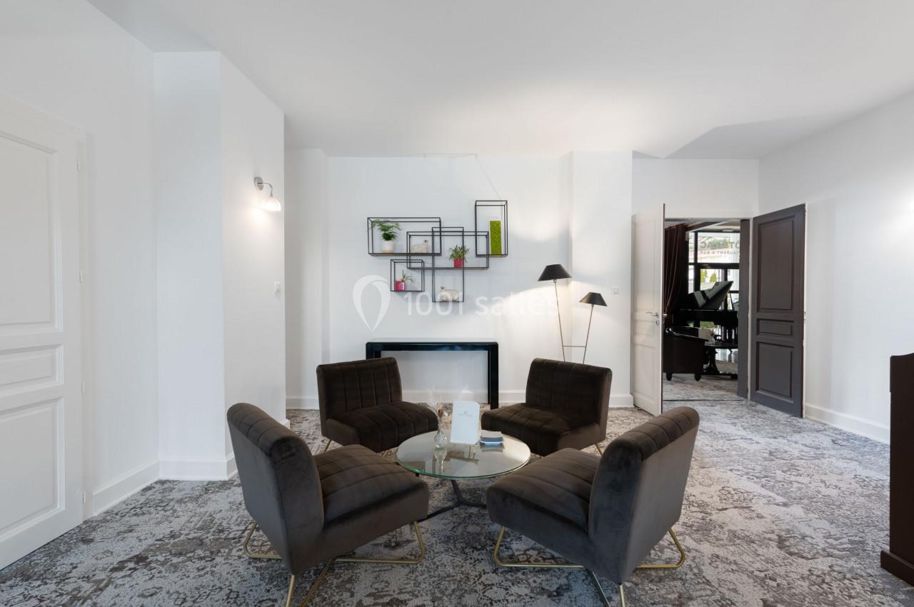 Salon lumineux avec quatre fauteuils gris autour d'une table basse en verre, étagères murales et piano visible au fond.