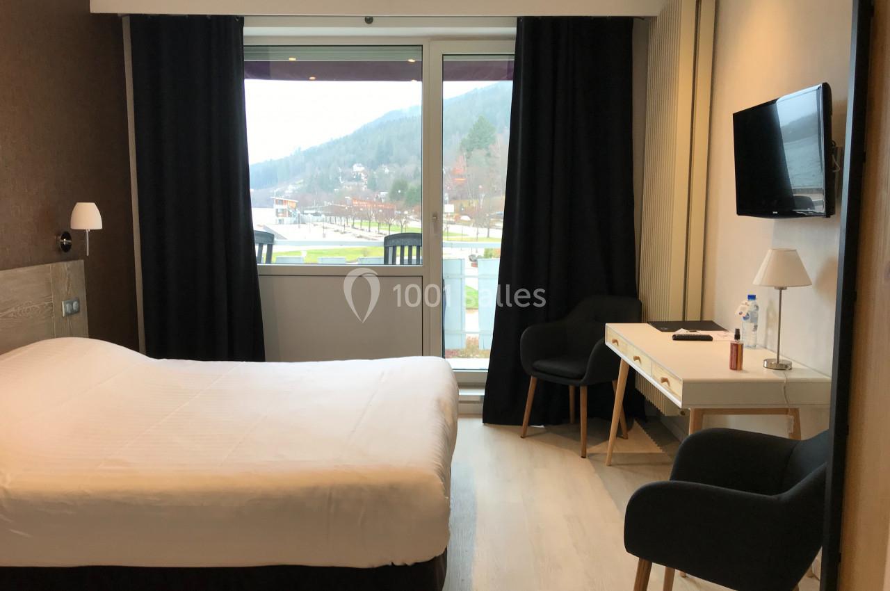 Chambre d'hôtel lumineuse avec lit double, bureau, deux fauteuils, télévision murale et grande fenêtre donnant sur un…