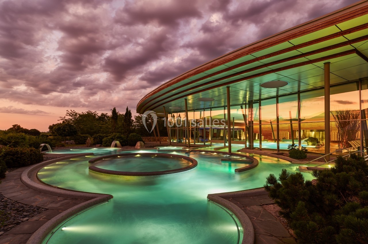 Piscine extérieure avec bassins circulaires illuminés au crépuscule, entourée de végétation et d'une architecture moderne.